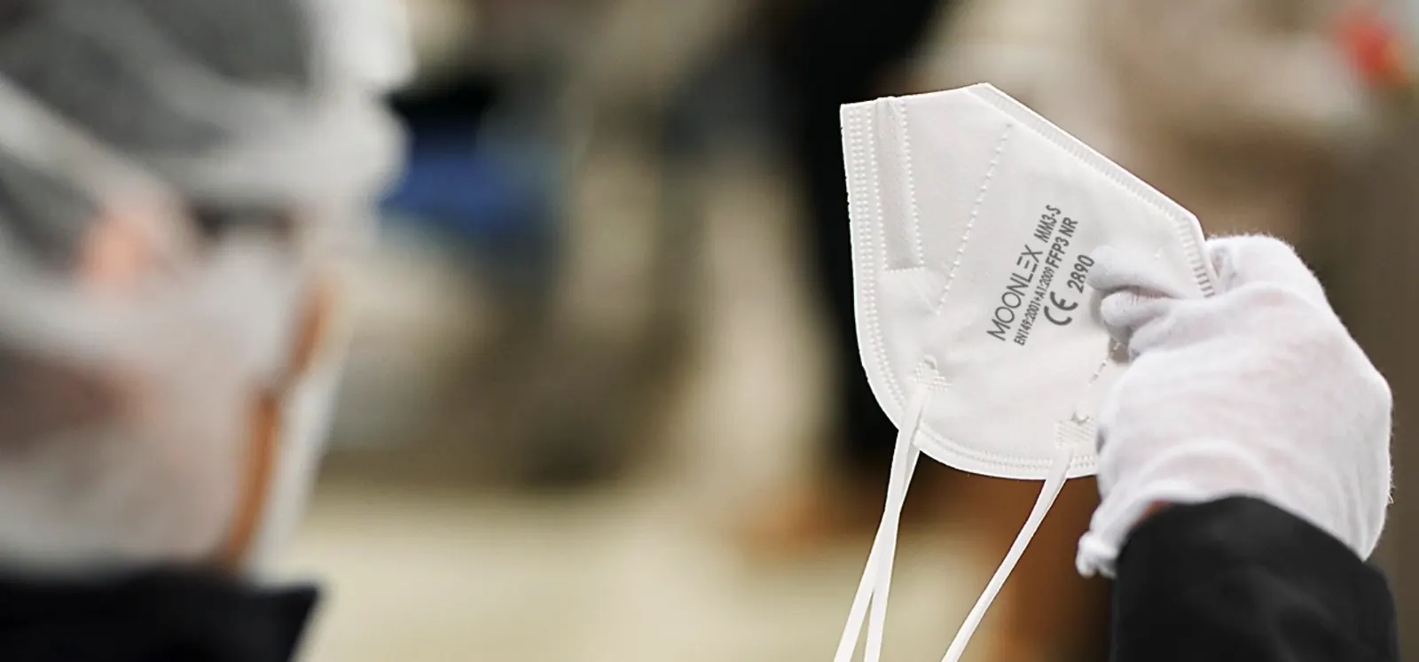 A gloved hand holding a folded MOONLEX respirator on the production floor